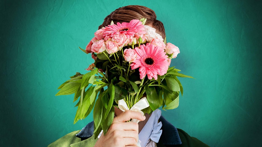 A man holds a bouquet of flowers in front of his face in a promotional image for The Importance of Being Earnest