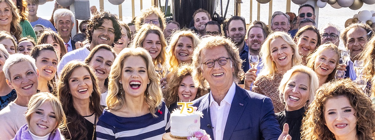 André Rieu stands alongside a birthday cake with a large group of people beside and behind him in a promotional image for André Rieu’s 75th Birthday Celebration.