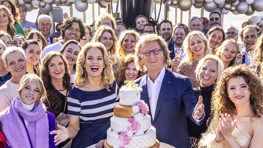 André Rieu stands alongside a birthday cake with a large group of people beside and behind him in a promotional image for André Rieu’s 75th Birthday Celebration.