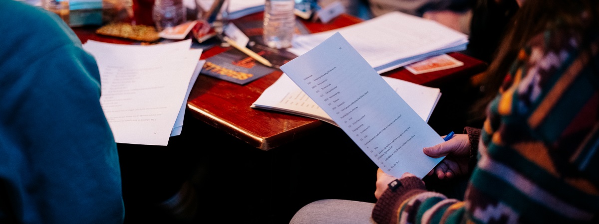 Someone holds a script with a desk in the background with various items including scripts, writing equipment, food and drink.