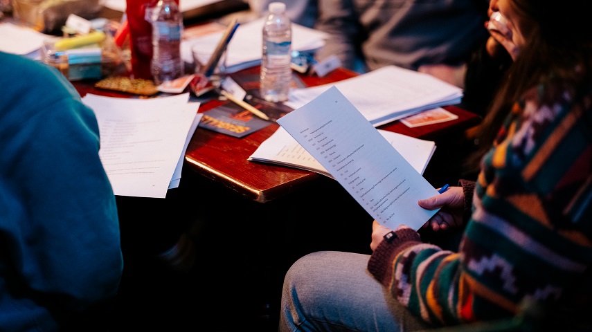 Someone holds a script with a desk in the background with various items including scripts, writing equipment, food and drink.
