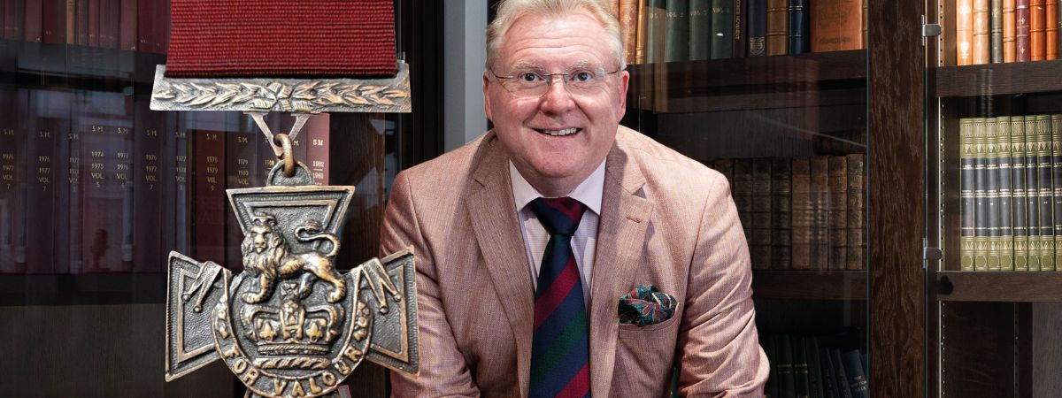 BBC's Antiques Roadshow's Mark Smith sits in a library space with an image of the Victoria Cross next to him.