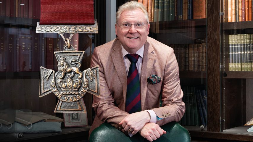 BBC's Antiques Roadshow's Mark Smith sits in a library space with an image of the Victoria Cross next to him.